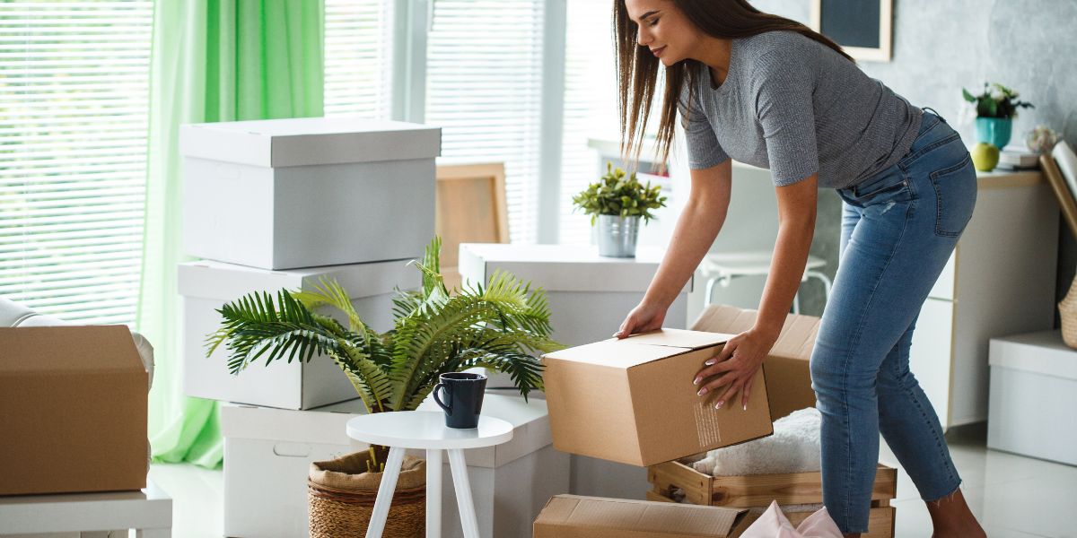 Woman packing a cardboard box in a living room filled with other boxes and plants, preparing for a home sale.